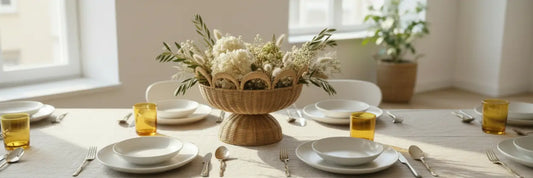 Tavola apparecchiata con stile: piatti in ceramica bianca, cestino decorativo in rattan Vanilla Fly come centrotavola, bicchieri ambra KnIndustrie Lime Line e posate Bamboo Bitossi Home con manico dorato